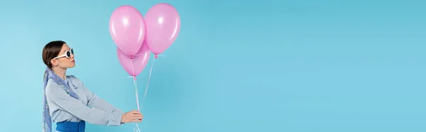 Mujer glamour en gafas de sol de pie con globos de color rosa sobre fondo azul, bandera - foto de stock