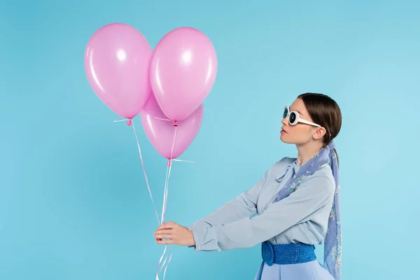 Молодая женщина в стильной одежде и солнцезащитных очках, держащих розовые шарики изолированы на синий — стоковое фото