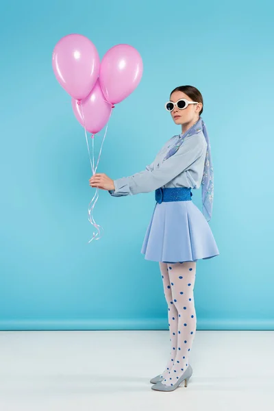 Модная женщина в солнечных очках и трико в горошек позирует с розовыми шариками на синем фоне — стоковое фото
