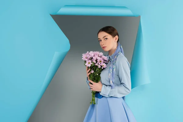 Trendy woman looking at camera while holding pink flowers near hole in blue paper on grey background — Stock Photo