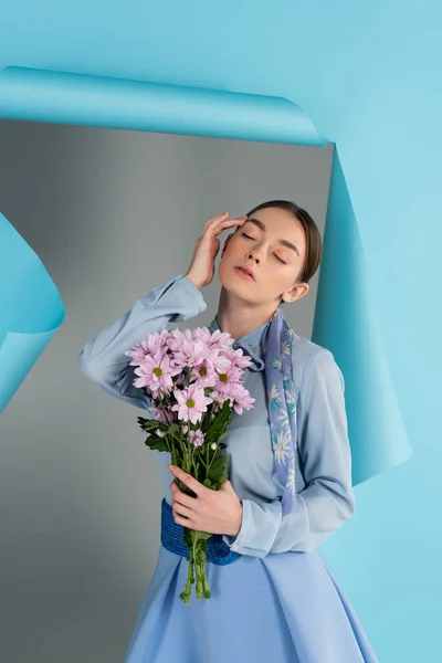 Елегантна жінка торкається обличчя, позуючи із закритими очима та квітами біля блакитного рваного паперу на сірому фоні — стокове фото