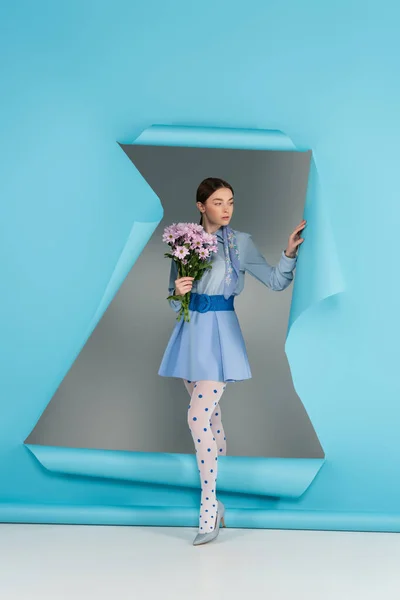 Trendy woman in dotted tights standing with pink flowers near hole in blue paper on grey background — Stock Photo
