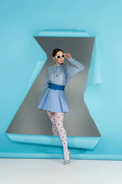 Giovane donna in abiti glamour regolazione occhiali da sole vicino foro in carta blu su sfondo grigio — Foto stock