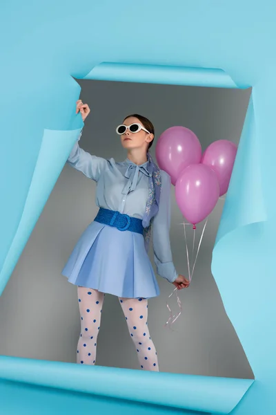 Гламурная женщина в трико горошек позирует с розовыми шариками возле отверстия в голубой бумаге на сером фоне — стоковое фото