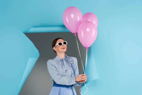 Счастливая модная женщина в очках, держащая розовые шарики рядом с голубой порванной бумагой на сером фоне — стоковое фото
