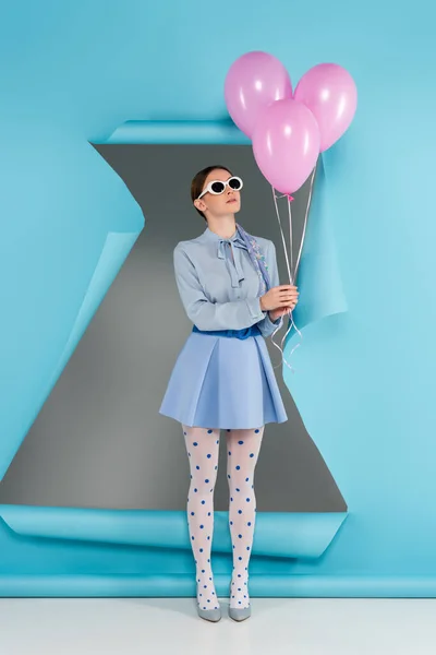 Модная женщина в точечных колготках и солнцезащитных очках позирует с розовыми шариками рядом с голубой порванной бумагой на сером фоне — стоковое фото