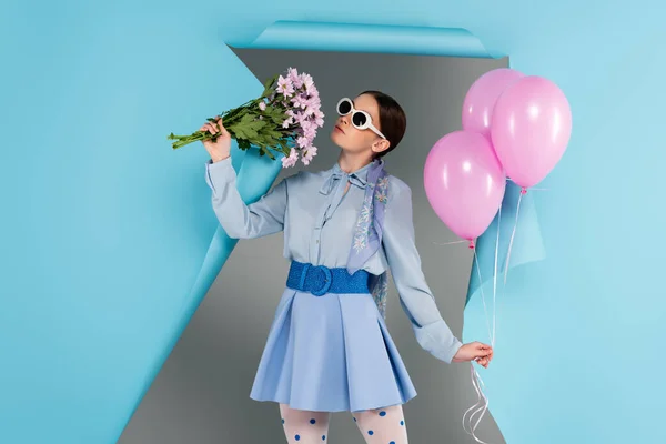 Стильная женщина позирует с розовыми цветами и воздушными шарами возле синей рваной бумаги на сером фоне — стоковое фото