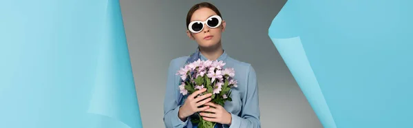 Гламурна жінка в сонцезахисних окулярах тримає рожеві квіти біля отвору в синьому папері на сірому фоні, банер — стокове фото