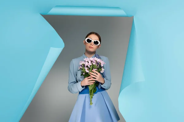 Модная женщина в солнечных очках, стоящая с розовыми хризантемы возле отверстия в голубой бумаге на сером фоне — стоковое фото