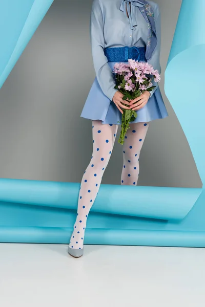 Vista recortada de la mujer glamour sosteniendo flores rosadas mientras sale agujero en papel azul sobre fondo gris - foto de stock
