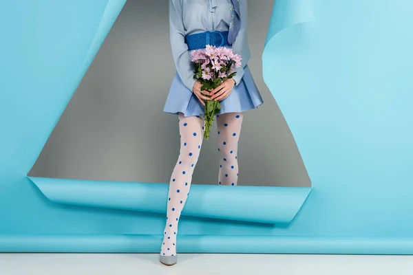 Vista parziale della donna glamour con crisantemi che esce dal buco in carta blu su sfondo grigio — Foto stock