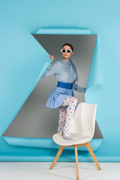 Elegant woman in dotted tights and sunglasses stepping on chair near blue ripped paper on grey background — Stock Photo