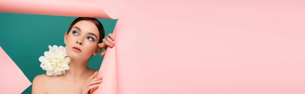 Mujer joven con peonía blanca en el hombro mirando hacia fuera agujero en papel rosa aislado en verde, bandera - foto de stock