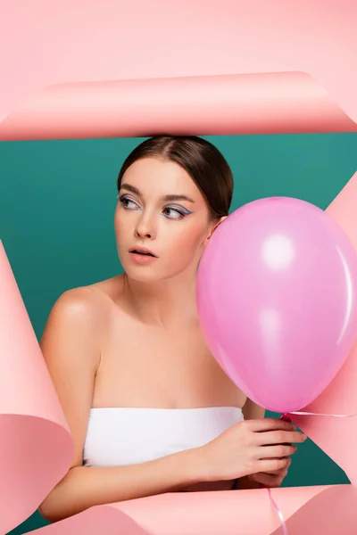 Bonita mujer con globos mirando hacia fuera agujero en papel rosa aislado en verde - foto de stock