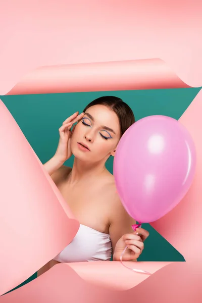 Young woman with closed eyes holding balloon near hole in pink paper isolated on green — Stock Photo