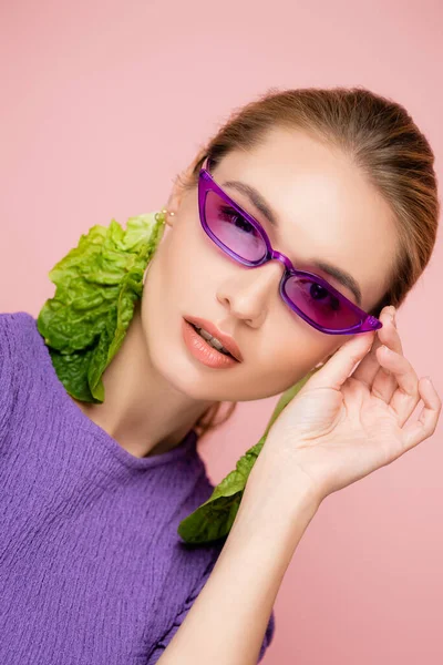 Femme élégante dans des boucles d'oreilles en laitue fraîche, touchant lunettes violettes isolées sur rose — Photo de stock