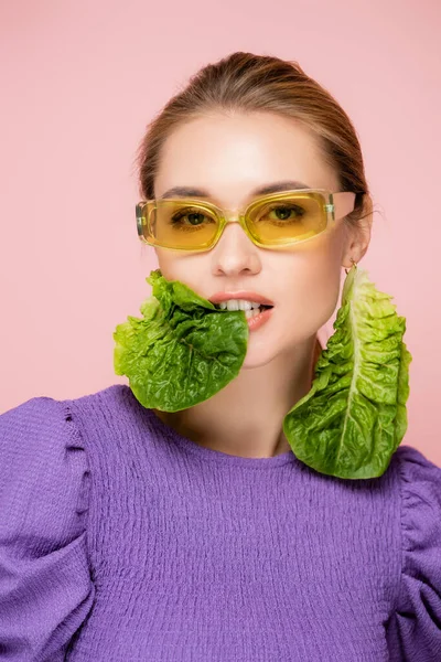 Femme sensuelle dans des lunettes de couleur, mordant boucle d'oreille de laitue fraîche isolé sur rose — Photo de stock