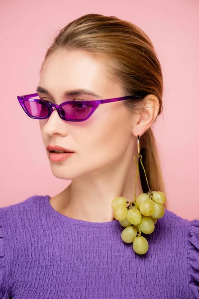 Bonita mujer joven en pendiente de uva madura y gafas púrpuras aisladas en rosa - foto de stock