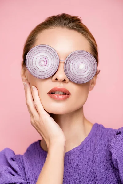 Young woman touching face while posing in eyeglasses with onion rings isolated on pink, surrealism concept — Stock Photo