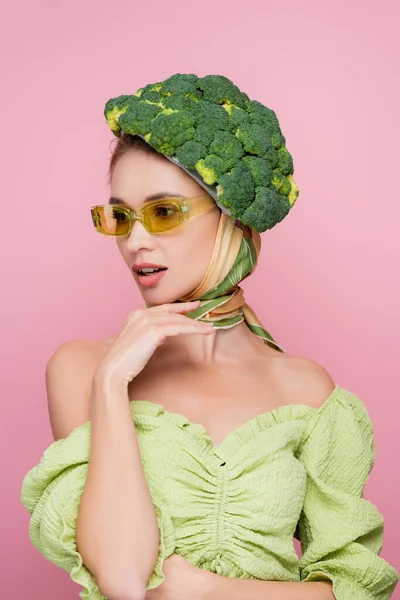 Mujer sensual en gafas de color y sombrero hecho de brócoli fresco aislado en rosa, concepto de surrealismo - foto de stock