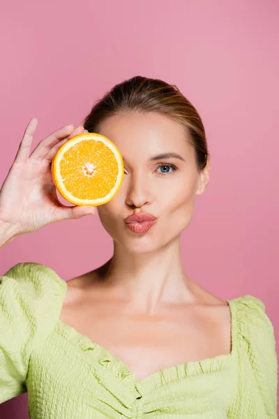 Bonita mujer enviando aire beso mientras cubre ojo con naranja mitad en rosa - foto de stock