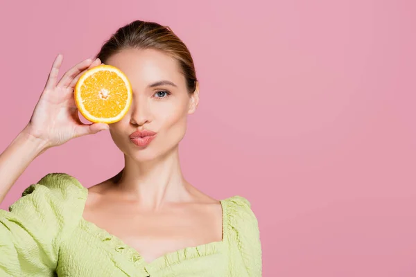 Sensual woman covering eye with half of juicy orange and sending air kiss isolated on pink — Stock Photo