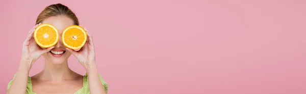 Молодая улыбающаяся женщина закрывает глаза половинками свежего апельсина, изолированного на розовый, баннер — стоковое фото