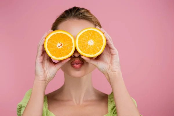 Joven mujer enviando aire beso mientras cubre los ojos con naranja mitades aisladas en rosa - foto de stock