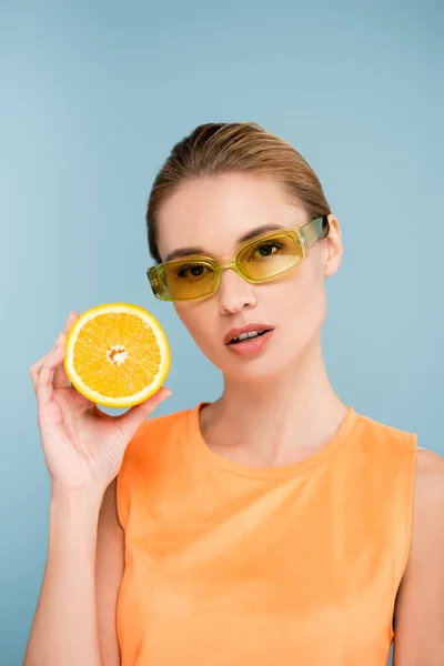 Молодая женщина в цветных очках держит половину сочного апельсина изолированы на синий — стоковое фото