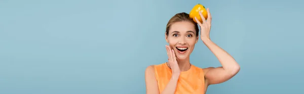Astonished woman holding yellow bell pepper on head while touching face isolated on blue, banner — Stock Photo
