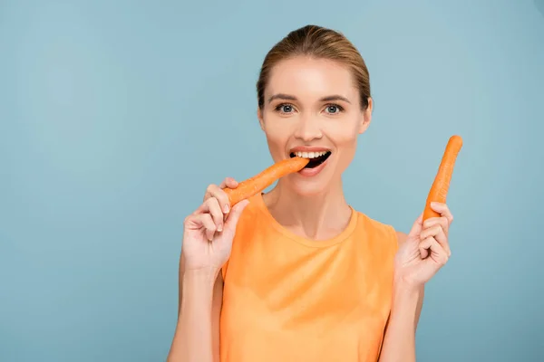 Pleased woman looking at camera while eating whole carrot isolated on blue — Stock Photo