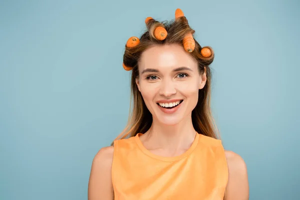 Femme joyeuse regardant la caméra tout en utilisant des carottes au lieu de bigoudis isolés sur bleu — Photo de stock