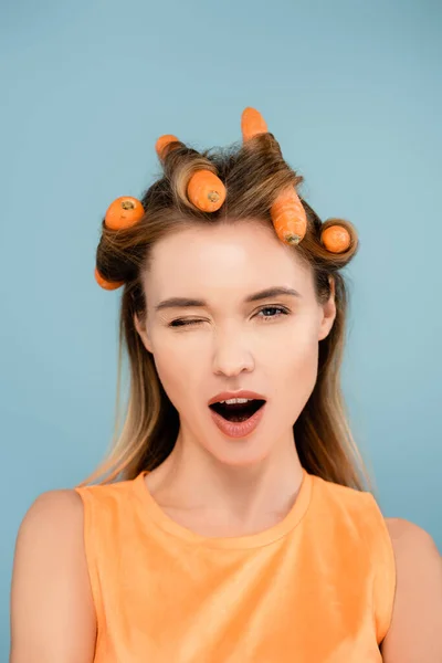 Flirty woman with carrots instead of curlers winking at camera isolated on blue — Stock Photo