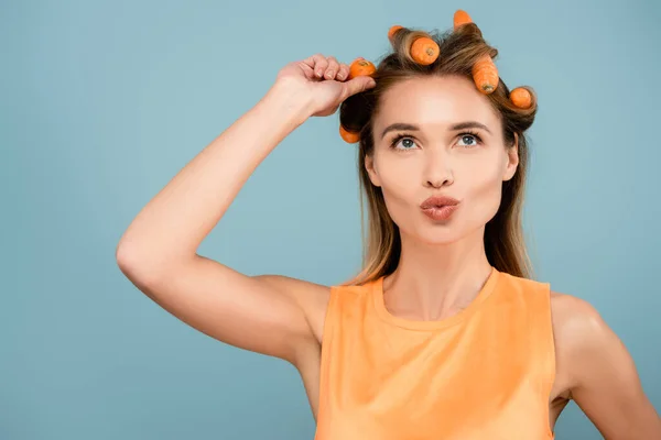 Cheerful woman pouting lips while waving hair with fresh carrots isolated on blue — Stock Photo