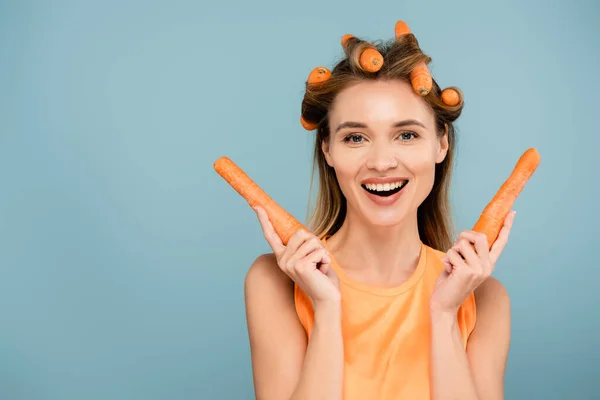 Jeune femme heureuse regardant la caméra tout en utilisant des carottes au lieu de bigoudis isolés sur bleu — Photo de stock