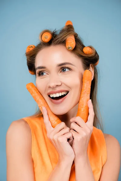 Mujer alegre mirando hacia otro lado mientras riza el cabello con zanahorias frescas aisladas en azul - foto de stock