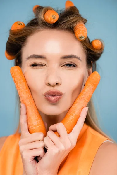 Femme gaie avec des carottes au lieu de bigoudis clin d'oeil et envoyer baiser d'air isolé sur bleu — Photo de stock