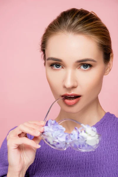 Mujer joven con maquillaje natural sosteniendo anteojos con flores cerca de labios aislados en rosa - foto de stock