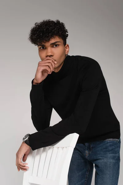 Giovane uomo africano americano in maglione dolcevita posa vicino sedia isolata su grigio — Foto stock