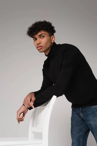 Young african american man in turtleneck sweater looking at camera and leaning on chair isolated on grey — Stock Photo