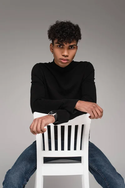 Curly african american man in turtleneck sweater sitting on chair while posing isolated on grey — Stock Photo