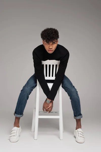 Full length of pierced african american man in turtleneck sweater leaning on white chair on grey — Stock Photo