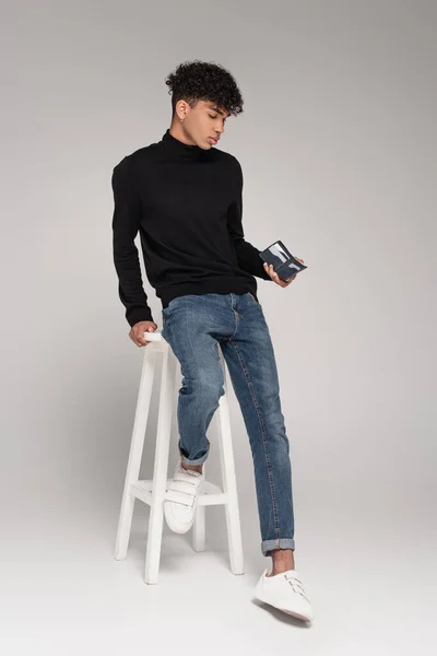Повнометражний молодий афроамериканський чоловік тримає шкіряний гаманець, спираючись на біле крісло на сірий — стокове фото