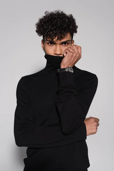 Curly african american man covering face with collar of black turtleneck sweater isolated on grey — Stock Photo