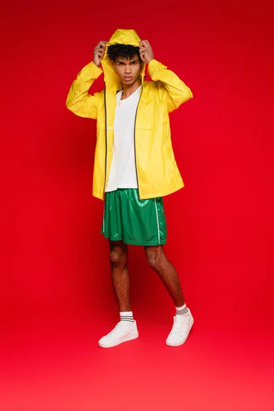 Full length of stylish african american man in yellow rain jacket adjusting hood on red — Stock Photo