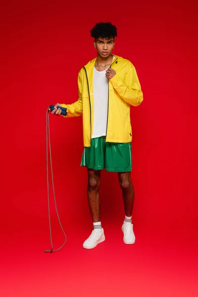 Full length of sportive african american man in yellow rain jacket holding jumping rope on red — Stock Photo