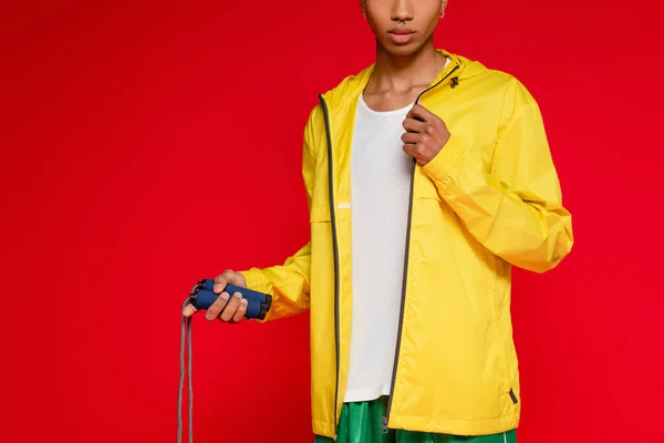 Cropped view of sportive african american man in yellow rain jacket holding jumping rope isolated on red — Stock Photo