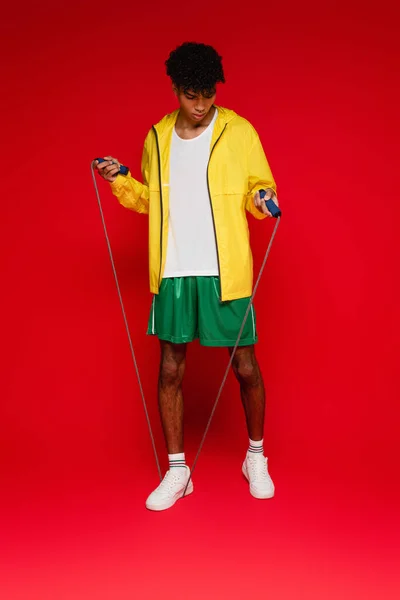 Full length of sportive african american man in yellow rain jacket exercising with jumping rope on red — Stock Photo