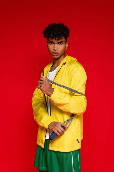 Pierced african american man in yellow jacket holding jumping rope isolated on red — Stock Photo