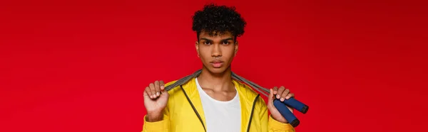 Perfurado afro-americano homem em casaco amarelo segurando pulando corda isolada em vermelho, banner — Fotografia de Stock